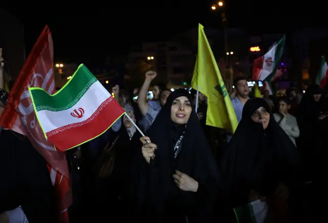 A woman, dressed in black, waves an Iranian flag. She is surrounded by others cheering an carrying flags.