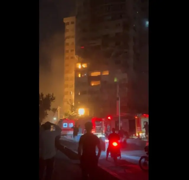 People stand in the street looking at a flaming high-rise building at night