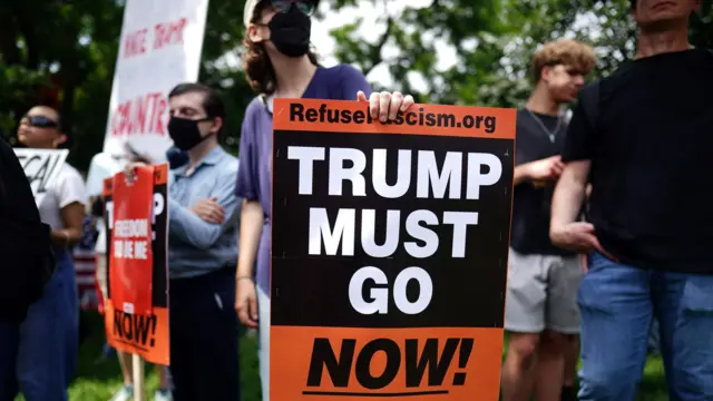 A man holds an orange and black sign that says "Trump must go now".