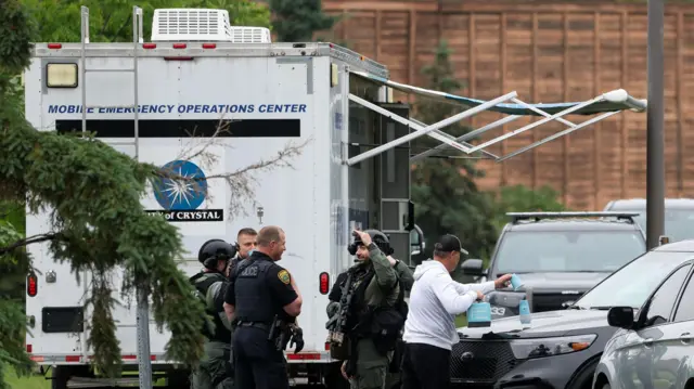Police officers, some armed with assault rifles, stand by some cars and in front of a mobile emergency operations centre which looks a bit like a white caravan