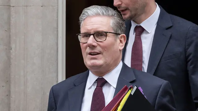 Mid-shot of Prime Minister Keir Starmer in black suit, white shirt and burgundy tie walking out of Number 10, Downing Street carrying a burgundy and black folder, a man in a matching suit and tie whose face is partially cut off walking behind him