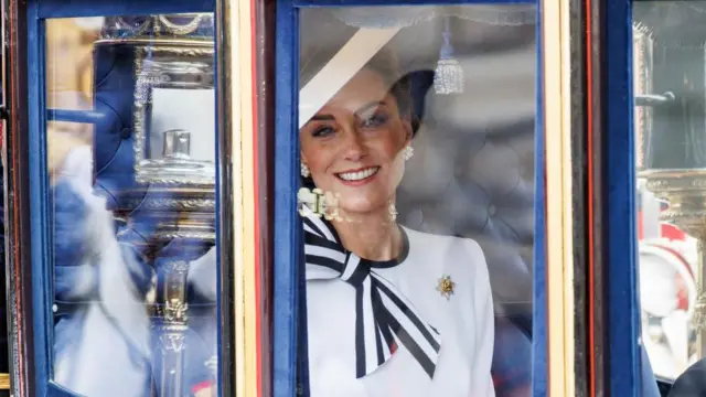 Catherine Princess of Wales smiles as she travels from Buckingham Palace to Horse Guards Parade inside a carriage during the Trooping the Colour parade in London in 2024. She smiles, wearing a white and blue outfit and hat.