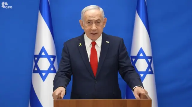 Benjamin Netanyahu standing behind a wooden podium, his hands grasping the sides. He's wearing a dark blue suit, white shirt and red tie, a blue background and two Israeli flag on each side of him