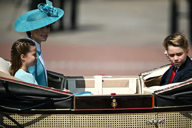 Catherine, Princess of Wales, with her children George, Charlotte and Louis in formalwear sit in a open carriage.