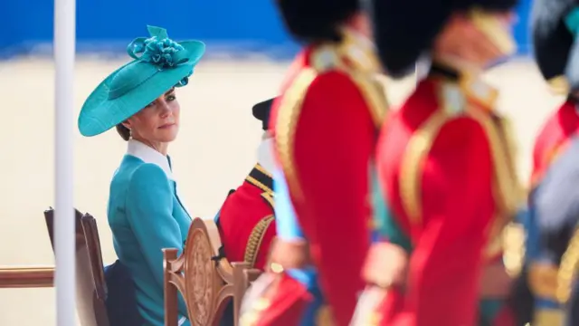 Catherine, Princess of Wales, attends the Trooping the Colour parade and wears a green jacket and hat. She looks behind a line of soldiers.