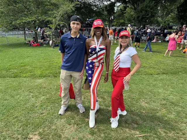 Two women and one man pose for a photo. The women are wearing red 'make America Great Again' hats. The woman in the middle is wearing a shirt and pants made out of the US flag.