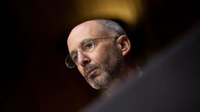 Close up photo of former US envoy Robert Malley (middle aged man with salt and pepper beard and air) and glasses sitting down in a dark room, he's he only illuminated bit in the frame