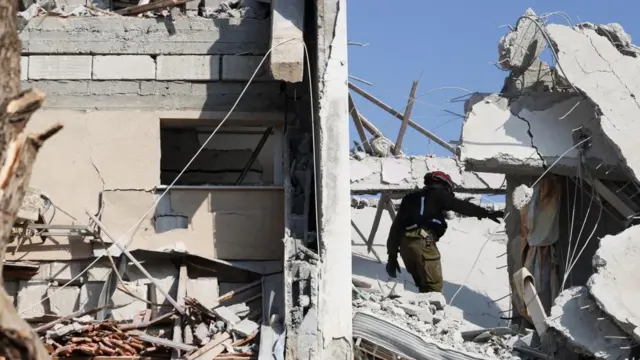 a rescue worker wearing a black jumper and cargo trousers and a red hard hat inspects damage. he is standing in the rubble of what appears to be an apartment block. concrete and rebar surround him.