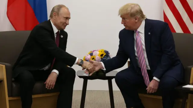 Vladimir Putin (L) sitting down in a black leather chair wearing a black suit shakes hands with Donald Trump (R) sitting on a matching chair in a dark blue suit, a wooden table with a vase of flowers on top sitting between them