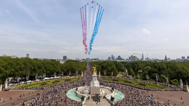 The Red Arrows, flanking an envoy of the RAF, approach Buckingham Palace during a flypast on the day of Trooping the Colour parade in 2023. Crowds of people stand outside Buckingham Palace and a RAF flyover zooms over them in the skies, projecting red, white and blue smoke.