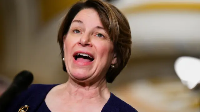 Amy Klobuchar, who is wearing a dark blue top and has short brown hair with pearl earrings