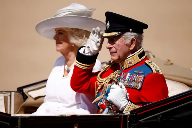 The King salutes wearing military uniform along Camilla in an open top carriage.