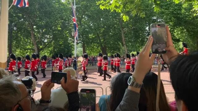Lines of soldiers march along the Mall with people watching and recording on their phones.