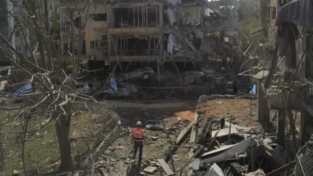 a worker in orange high-vis stands amid rubble at the site of a strike in tel aviv.