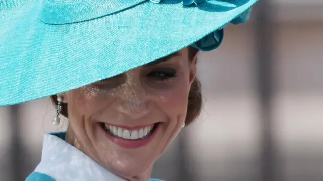 Catherine wears a green suit jacket and hat and smiles.