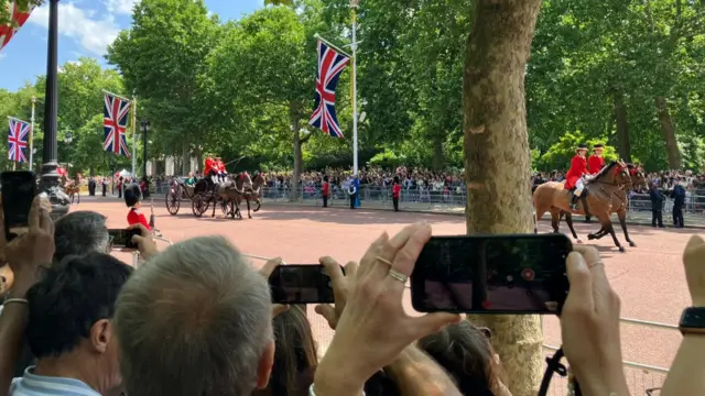 People hold their phone cameras high along the Mall trying to take pictures of a carriage going past.