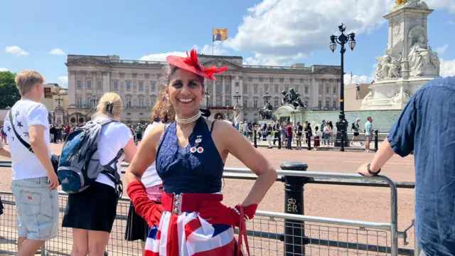 A woman stands outside Buckingham Palace with a red hat and gloves and a Union Jack skirt.