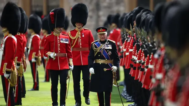 King Charles III inspects the regiment during a ceremony at Windsor Castle. He wears a black uniform surrounded by personnel in a red jacket and a long black cap welsh guard uniform.