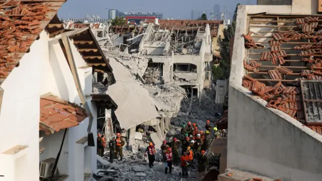 about twenty workers dressed in high vis and helmets work in the rubble of a residential building in tel aviv