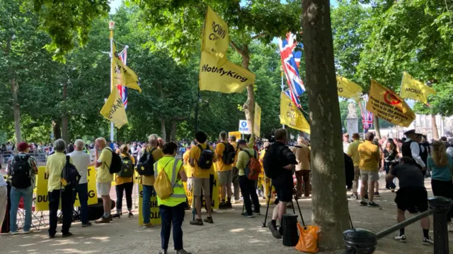 A group of people wave yellow flags near Buckingham Palace. One reads: 'Not my King'.