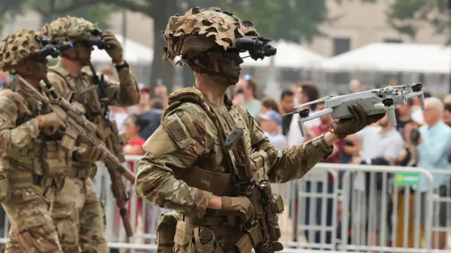 A solider with his face coated in camouflage paint and night-vision goggles on his helmet holds up a small drone. Two soldiers march behind him.