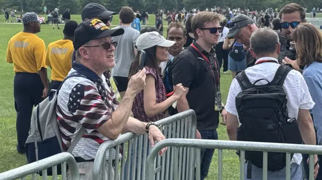 A man wearing an American flag shirt leans against a barricade