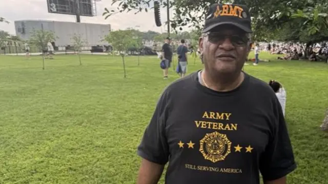 A man wearing a military hat and a shirt that says "Army Veteran" stands in a grassy area