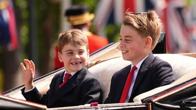 Louis smiles and waves alongside brother George in an open top carriage.