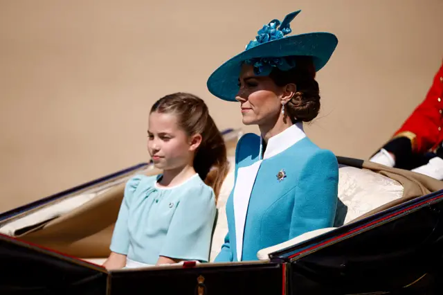 Catherine wears a green jacket and hat alongside Charlotte in an open top carriage.