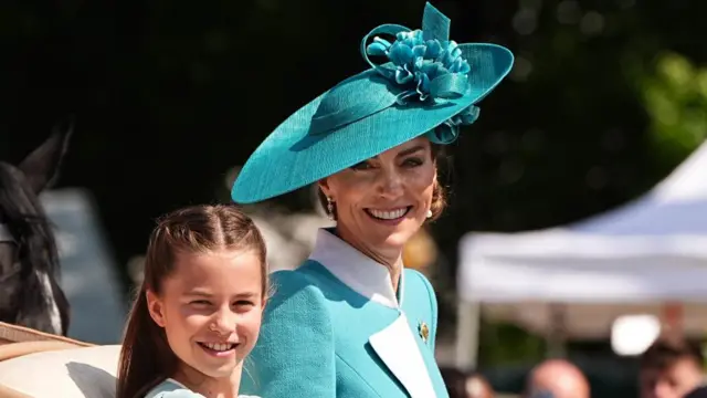 Catherine smiles wearing a green outfit and hat, sitting next to Charlotte who also smiles.