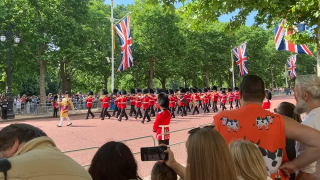 A group of soldiers play trumpets in red uniform and long black hats. They walk along the Mall.