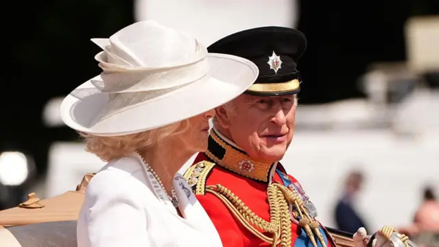 Charles and Camilla are sitting in a carriage. Charles wears a red uniform and black hat, Camilla wears a white outfit and hat.