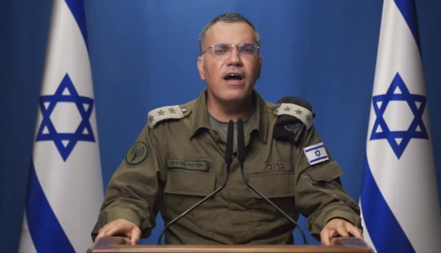 Avichay Adraee speaking in front of a microphone in green uniform, Israeli flags behind him.
