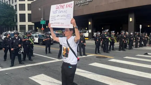 A man holds a sign over his head that reads "Deport kings not families" as police officers stand in a line behind him.