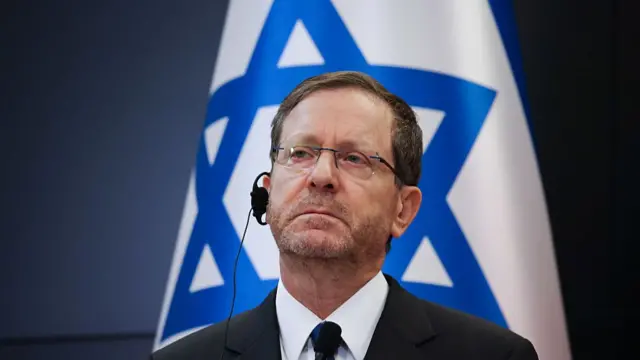Isaac Herzog looks on with a ear piece, wearing a suit and seated in front of an Israeli flag