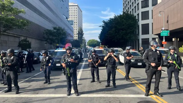 Police officers wearing helmets and face shields stand in a row in front of numerous police vehicles.