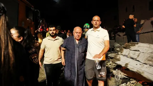An elderly man in dark blue nightgown behind escorted by two younger men wearing white and brown polo shirts. Surrounding them is debris.