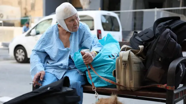 an old woman sits on a bench, her head wrapped in bandages. she is smoking a cigarette and holding a red lead for a dog. luggage is piled on the bench next to her