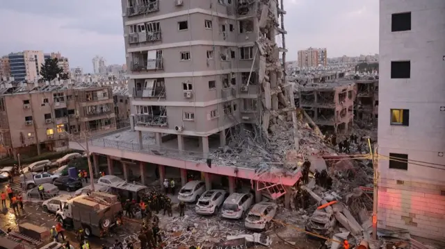 Wide shot of a damaged grey building. A crowd of first responders has gathered in front of it.