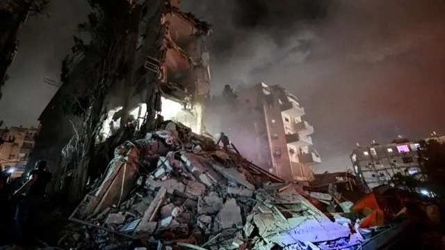 Wide shot of a severely damaged building in the dark, illuminated by lights from first responders.