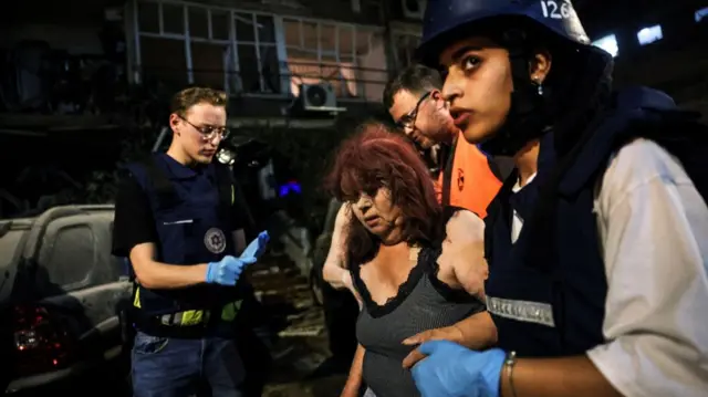 An injured middle-aged woman in a tank top being escorted by another woman with a helmet and black vest.