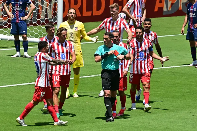 The referee puts his red card back in his pocket as Atletico players protest
