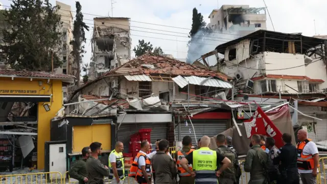Group of people stood in front of a damaged building, debris on roof and floor, smoke rising in background.