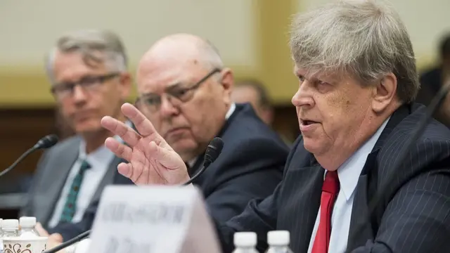 A man gestures while speaking behind a microphone on a panel hearing.