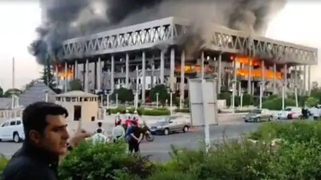 Man standing outisde pointing at the IRIB building on fire.