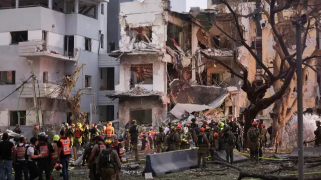 Heavy damage to a building, with dozens of rescue workers standing near it.