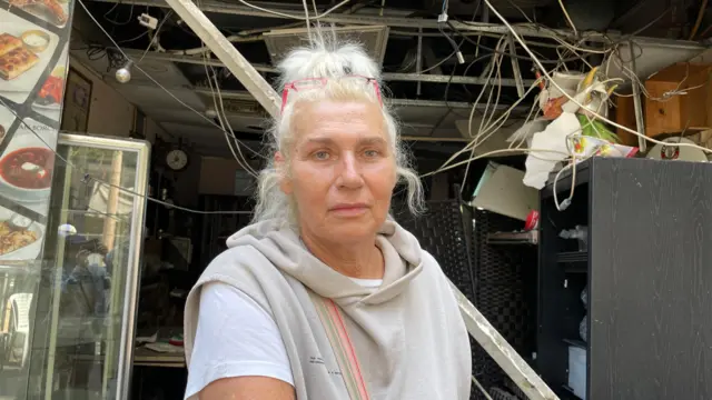 A woman with blonde hair and glasses on her head, stood infront of a damaged bar