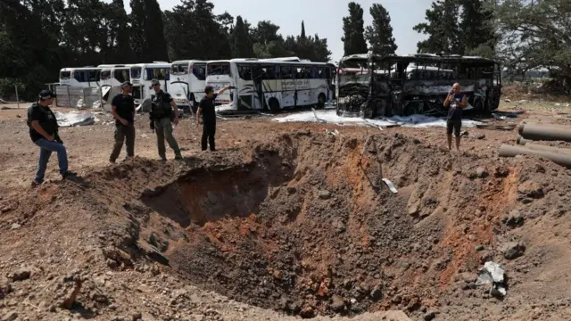 People stand near a crater in the ground with parked buses behind them