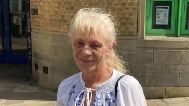 Amanda Bassett smiles at the camera while standing outside Luton Crown Court. She has long grey hair that is tied back and she wears a white and blue blouse.