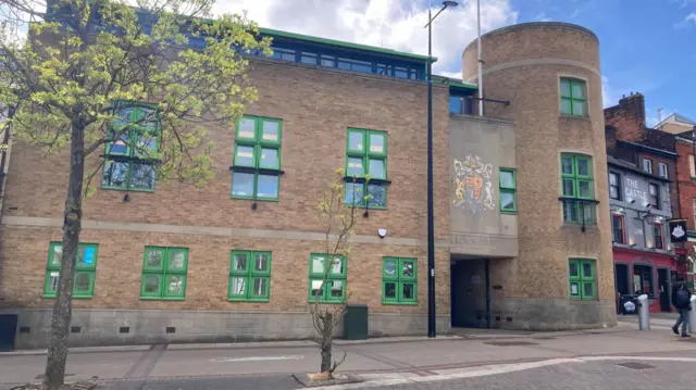 Exterior of Luton Crown Court. A three-storey, modern, pale brick building with green-framed windows. The front features a rectangular block and a round tower either side of the main entrance